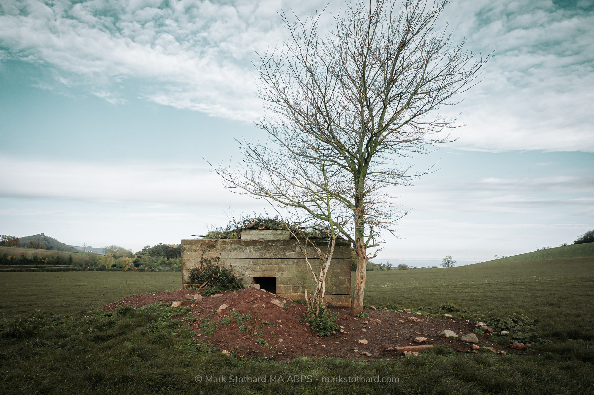 A Second World War pillbox near Carhampton, west of Minehead, remains remarkably well-preserved, offering a tangible reminder of Britain&rsquo;s wartime defences.Constructed of concrete in a hexagonal design, this pillbox originally sat nestled in the corner of a field. However, the removal of hedgerows in 1994 has left it standing exposed in the middle of the field, with its door facing northeast. Aerial imagery suggests the structure was strategically positioned to face north, with its entrance likely located on the south side.The pillbox features an unusual, non-standard design. Inside, a concrete step, approximately 1 foot high, leads to a raised platform measuring 3 feet by 3 feet and standing 2 feet high. The entrance is notably low, at just 3 feet in height, a feature typical of such defensive structures. An internal ricochet wall further demonstrates its defensive purpose, designed to minimize the risk of direct fire reaching occupants. Interestingly, part of the roof appears to be metal, supported by wooden rafters, with wood cladding applied externally. Historical records indicate it was once camouflaged by a hedge, blending it into the rural landscape to obscure its presence from potential attackers.