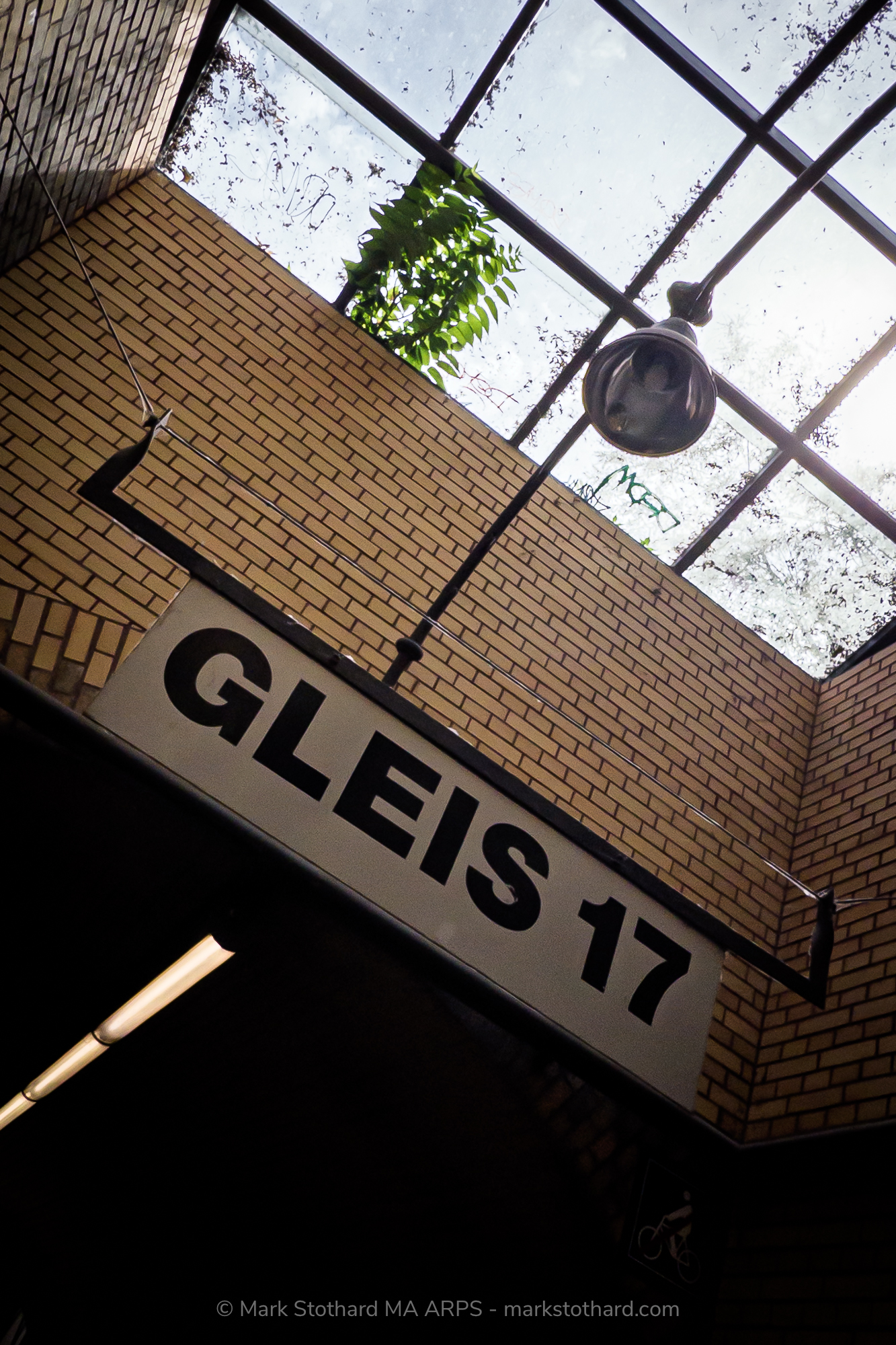 An upward view of the Gleis 17 sign at Platform 17 in Berlin. Brick walls rise toward a glass roof where weeds push through cracked panes. A single lamp hangs above. This was the departure point for thousands of Jews deported during the Second World War to concentration camps and death.
