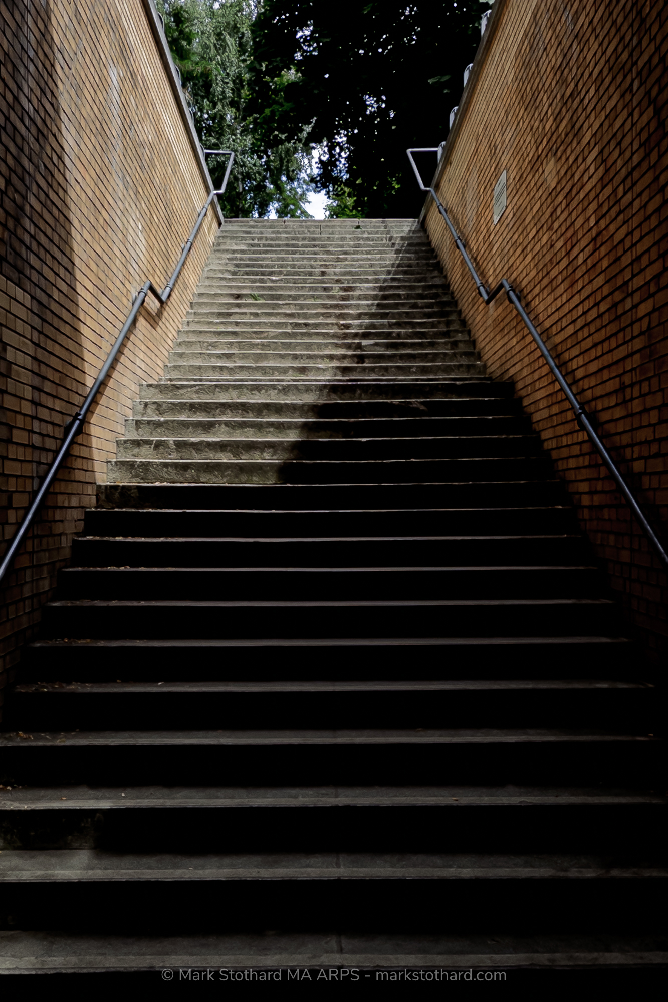 A flight of concrete steps rises between brick walls at Platform 17 in Berlin. Light falls across the stairway, dividing shadow from brightness. This quiet exit stands above the tracks from which thousands of Jewish men, women, and children were deported during the Second World War.