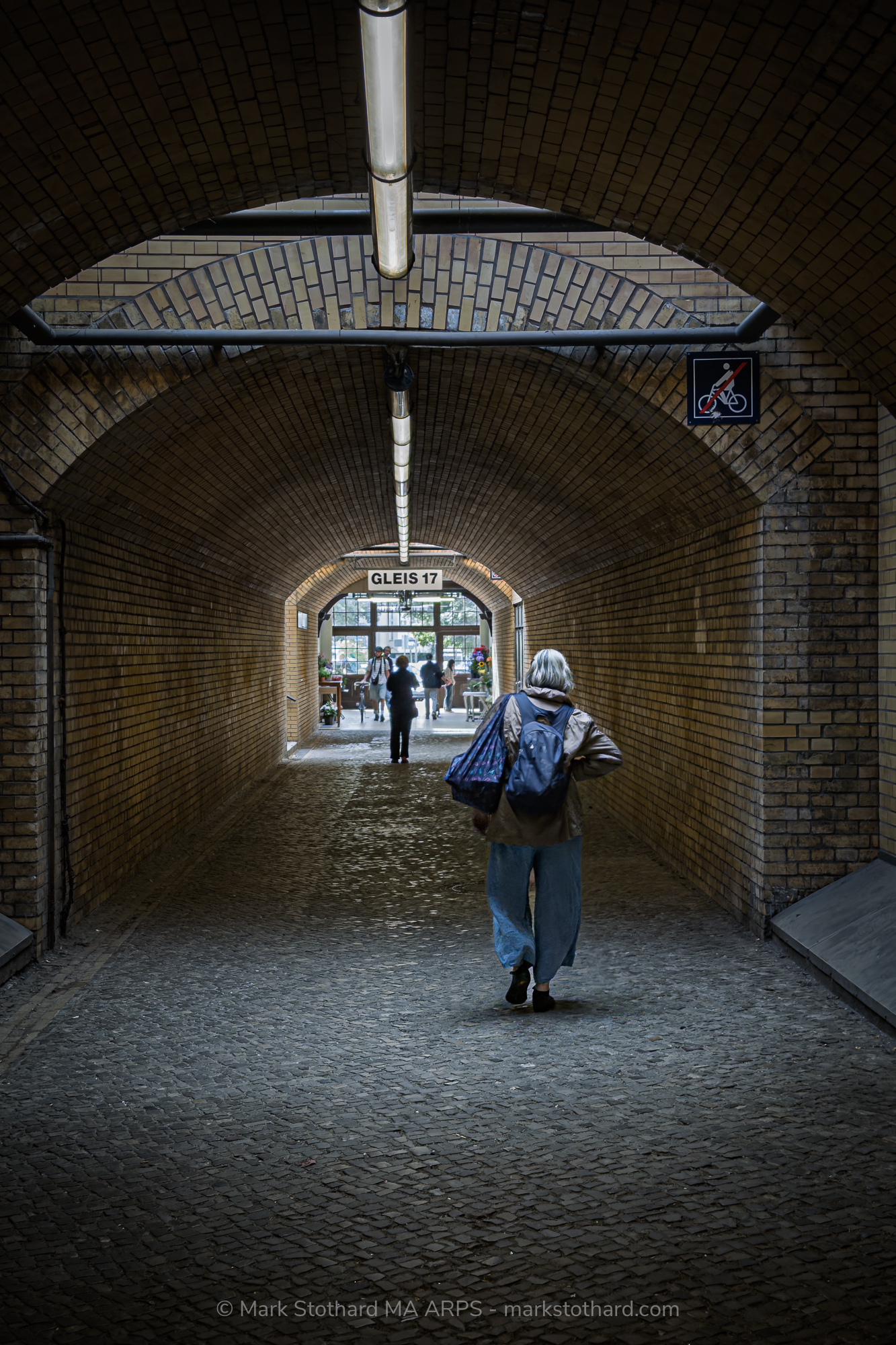 This photograph shows the brick underpass beneath Platform 17 at Grunewald station in Berlin. A single figure walks away from the camera, carrying a bag and wearing a backpack. The arched ceiling and repeating brickwork create strong symmetry. Overhead lights run along the centre line, guiding the viewer&rsquo;s gaze toward the bright opening at the far end.A sign reading Gleis 17 hangs above the exit. Today, commuters and visitors pass through this space without pause. During the Second World War, Jews were forced through these platforms and loaded onto trains bound for concentration camps. This underpass formed part of that route.The image holds tension between past and present. The bricks remain. The structure remains. Daily life continues. Yet the location carries the memory of deportation and loss. The solitary figure becomes a quiet witness, walking through a corridor shaped by history.