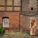 A somber, documentary-style photograph capturing the weathered, industrial brick facade at Platform 17 (Gleis 17) at Berlin-Grunewald Station, Germany. This site holds profound historical significance as a major deportation point for Jewish citizens during the Holocaust under the Nazi regime. The image focuses on the textures of memory, featuring a heavily rusted metal door, a sealed window, and aged brickwork, juxtaposed with the subtle encroachment of green vegetation and the remnants of gravel railway ballast. The composition stands as a quiet, haunting witness to the trauma associated with this place of memory and historical reckoning.