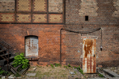 A somber, documentary-style photograph capturing the weathered, industrial brick facade at Platform 17 (Gleis 17) at Berlin-Grunewald Station, Germany. This site holds profound historical significance as a major deportation point for Jewish citizens during the Holocaust under the Nazi regime. The image focuses on the textures of memory, featuring a heavily rusted metal door, a sealed window, and aged brickwork, juxtaposed with the subtle encroachment of green vegetation and the remnants of gravel railway ballast. The composition stands as a quiet, haunting witness to the trauma associated with this place of memory and historical reckoning.