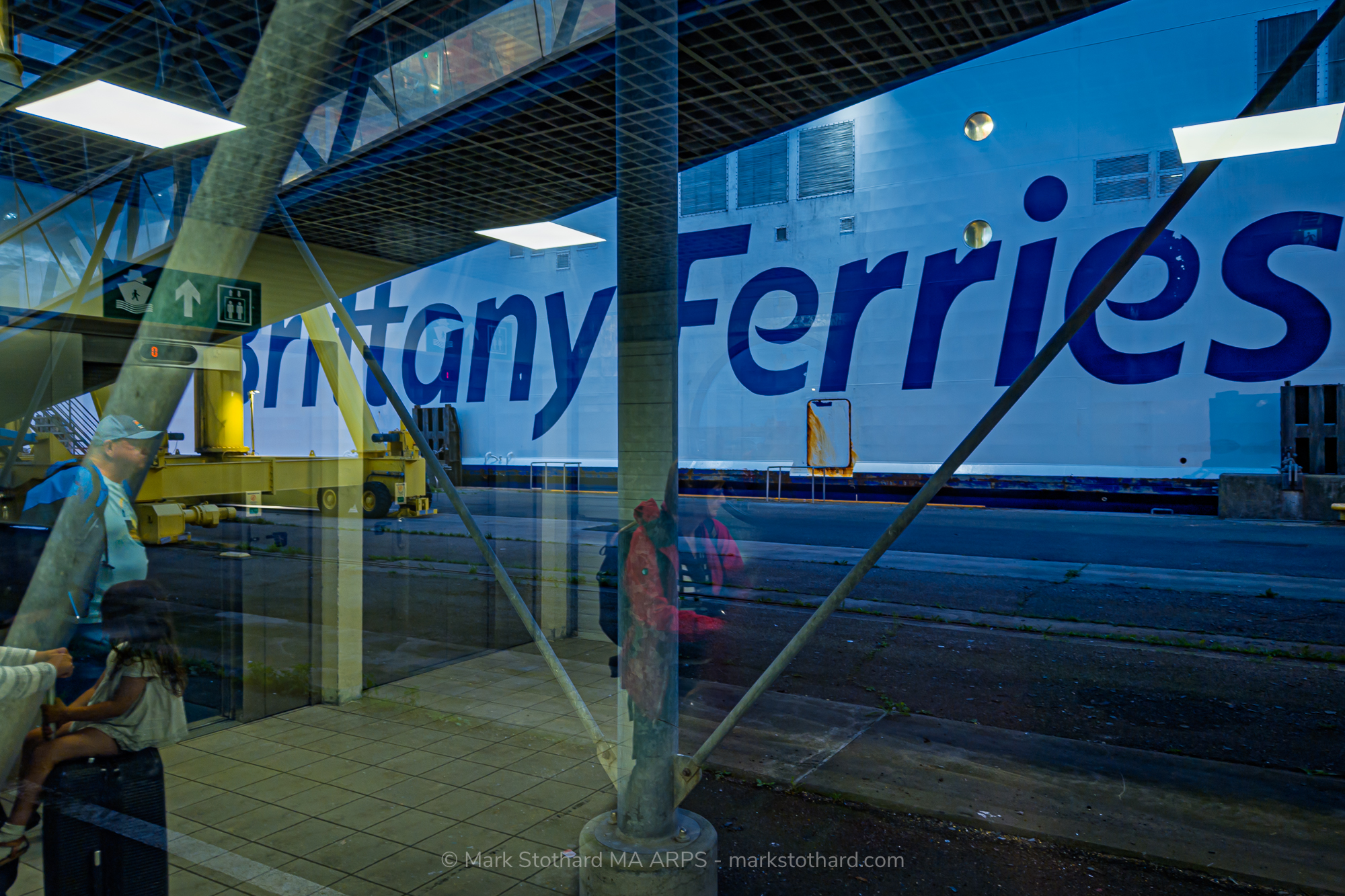 Passengers wait within the glass corridor of the Ouistreham ferry terminal as the vast hull of Brittany Ferries Mont St Michel looms outside. The scene becomes a layered reflection of the journey and pause, with travellers’ silhouettes merging with the industrial geometry of beams, lights, and the ship’s colossal lettering. Through the transparent barriers, reality and memory overlap: the promise of landfall against the lingering rhythm of the sea.
