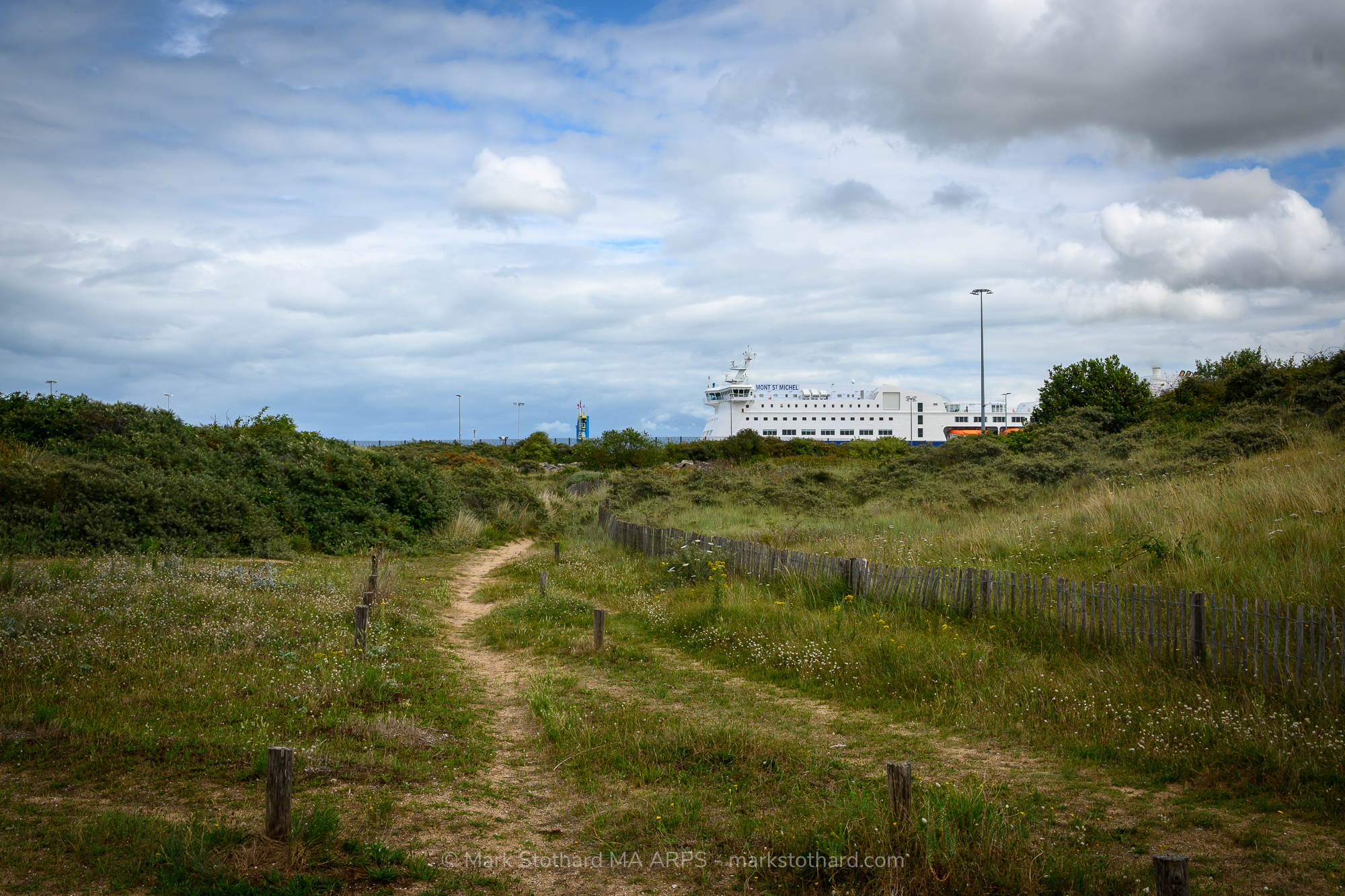 A winding coastal path leads through the wild grasslands of Ouistreham’s dunes, guiding the eye toward the towering Brittany Ferries vessel Mont St Michel. The white ship, poised for departure, stands out sharply against the green earth and the grey-blue sky above. The ferry’s presence introduces a human rhythm into this tranquil natural setting a moment caught between stillness and motion, land and sea, departure and return.