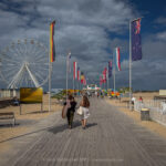 The promenade unfolds beneath drifting clouds in Ouistreham, France, a walkway lined with flags from across the world, whispering stories of unity and remembrance. The Ferris wheel stands silent, a circular echo of time and motion, while beach huts and distant waves recall both leisure and legacy. Sunlight flickers across the boards, where footsteps of locals and travellers intertwine, each carrying their own quiet narrative toward the horizon.
