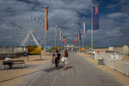 The promenade unfolds beneath drifting clouds in Ouistreham, France, a walkway lined with flags from across the world, whispering stories of unity and remembrance. The Ferris wheel stands silent, a circular echo of time and motion, while beach huts and distant waves recall both leisure and legacy. Sunlight flickers across the boards, where footsteps of locals and travellers intertwine, each carrying their own quiet narrative toward the horizon.