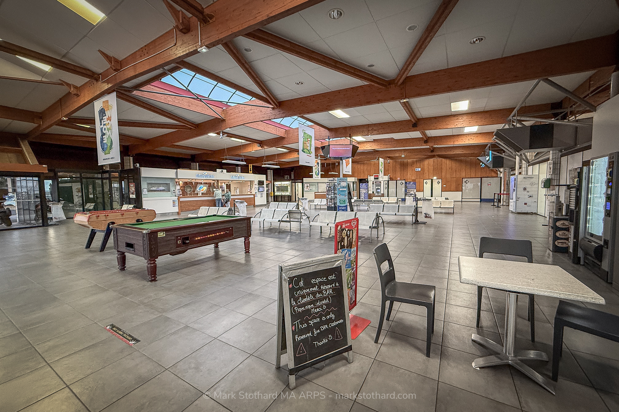Inside the Ouistreham Ferry Port terminal in Normandy, France, light spills gently through the skylights, illuminating a calm moment between journeys. The hall’s wooden beams and tiled floor frame an atmosphere of transience, where games of table football and pool stand ready to distract travellers from the slow rhythm of waiting. A chalkboard sign reserves a modest café space, a reminder of human order amid the ebb and flow of travel. Outside, ferries await their next crossing to England, while inside, time seems to pause, caught between departure and arrival.