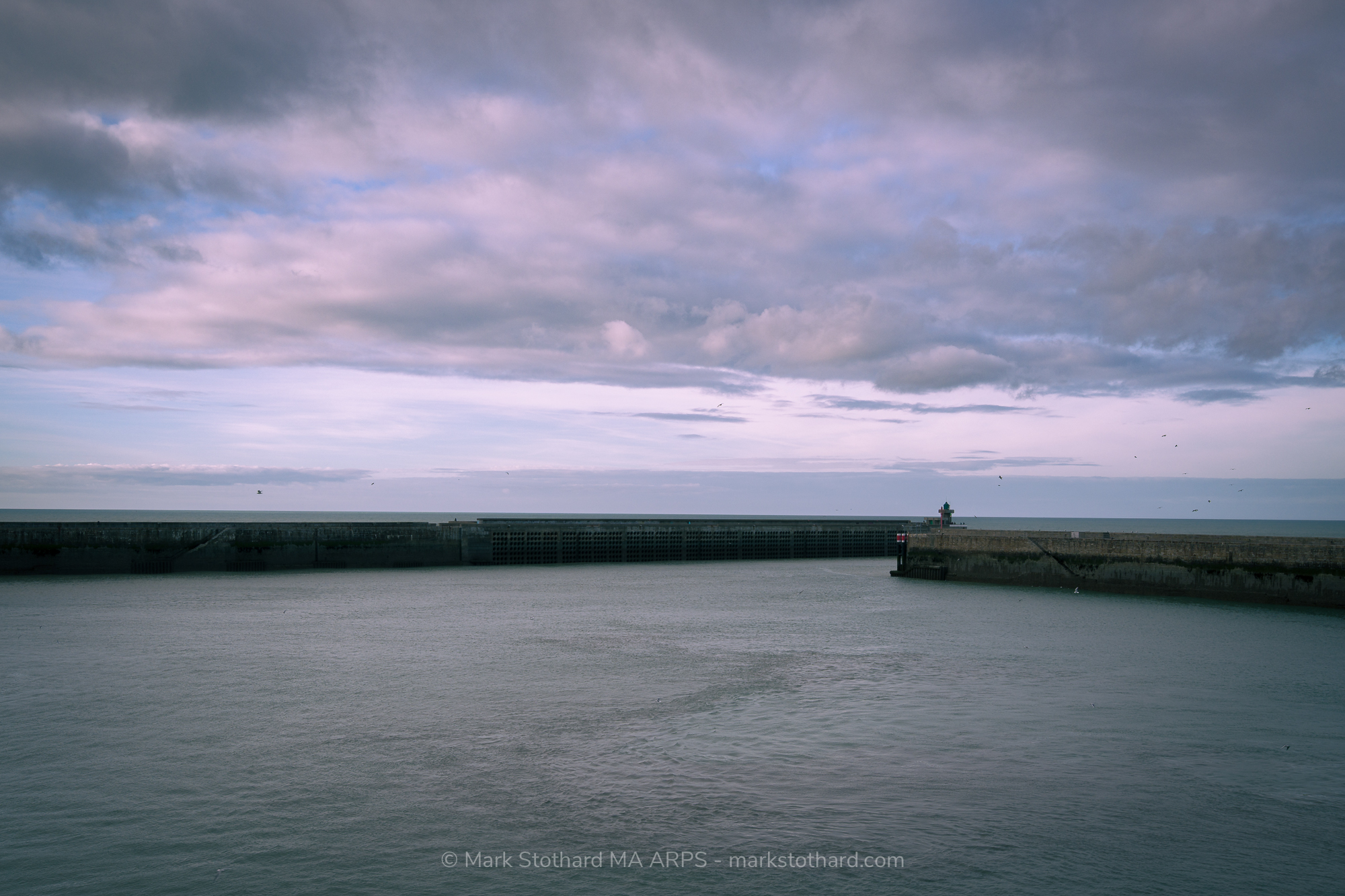 Dieppe harbour opens toward the English Channel under a wide afternoon sky. The long breakwater forms a hard line across calm water. A small lighthouse marks the exit. Beyond the wall lies the unseen coast of Britain. Gulls circle in cold air while clouds gather above the quiet port tonight.
