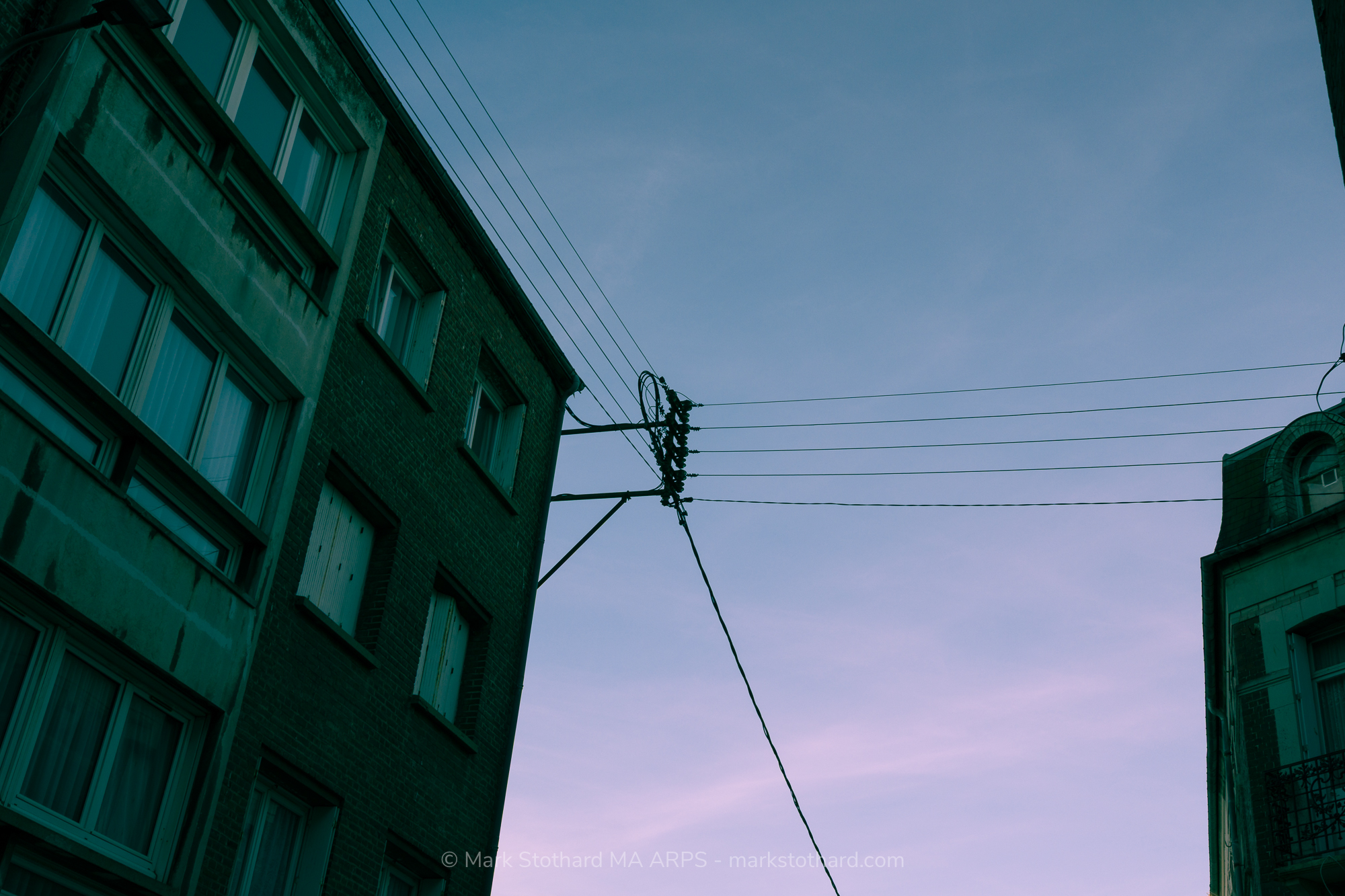 Standing in a narrow street in Dieppe the camera turns toward the sky. Old buildings lean inward. Power lines stretch and knot above the gap between roofs. Fading evening light softens the scene. The view feels quiet, enclosed, and slightly tense, as if the town holds its breath overhead tonight.