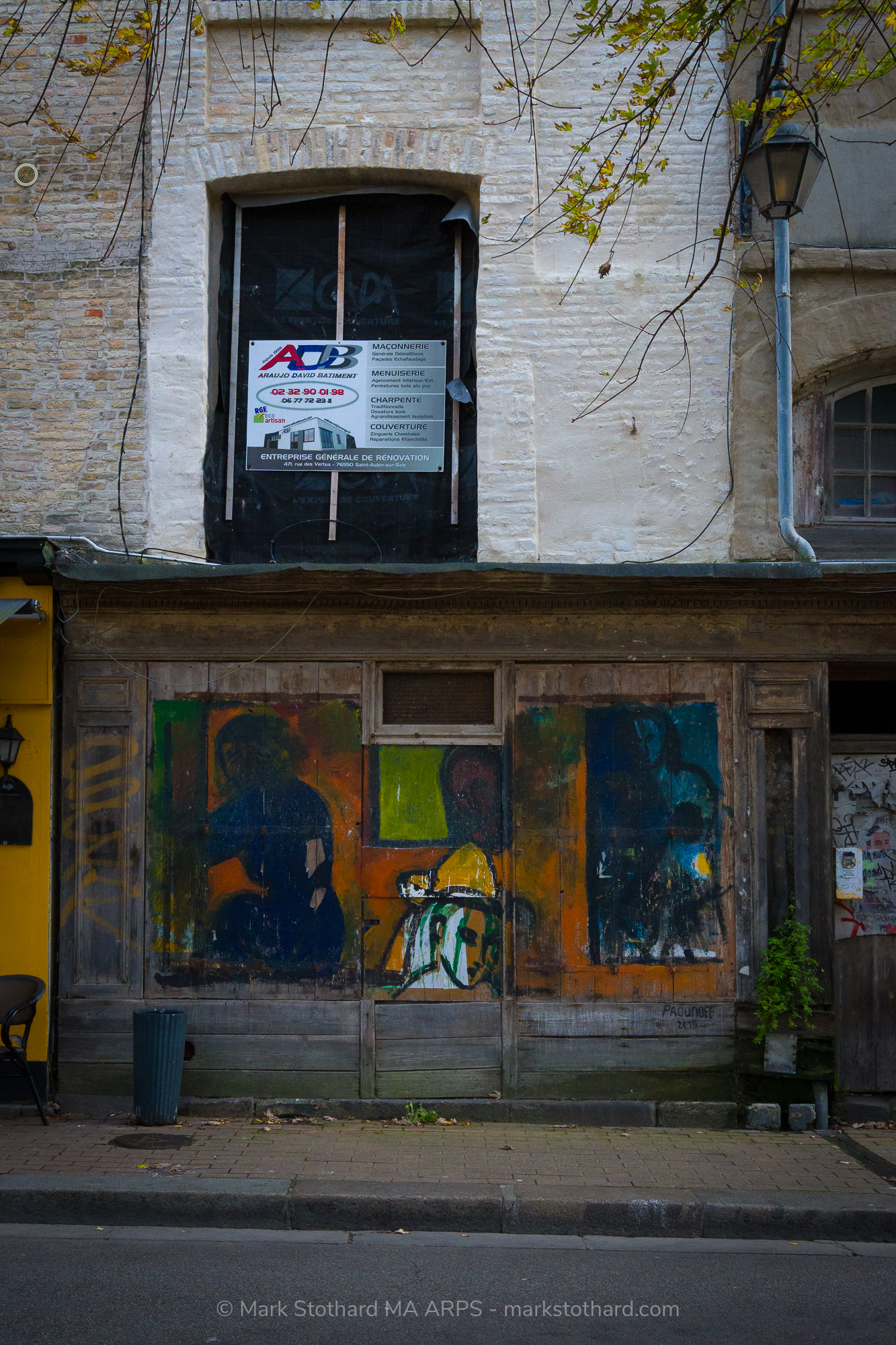 A boarded shopfront in Dieppe carries fading graffiti portraits across weathered wooden panels. Above, a sealed window and renovation notice signal suspended time. The street is quiet. Leaves hang from branches overhead. Art, decay, and waiting meet in one fa&ccedil;ade that records passing lives and unfinished stories