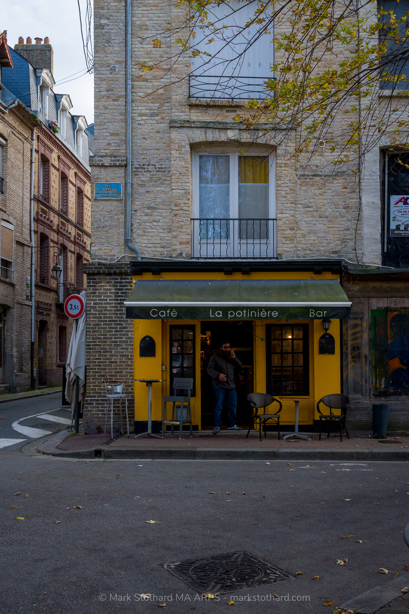 In a quiet corner of Dieppe a small yellow caf&eacute; sits beneath pale stone walls. A man pauses in the doorway while empty chairs wait on the pavement. Autumn leaves scatter across the street. The scene feels calm, ordinary and quietly human within the narrow streets of this harbour town.