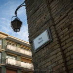 An old street sign reading Rue Denys sits fixed to a weathered brick corner in Dieppe. A traditional iron lamp hangs above the junction while a modern apartment block rises opposite. The image contrasts textures, eras, and urban change, revealing quiet layers of the town’s architecture and everyday geography today.