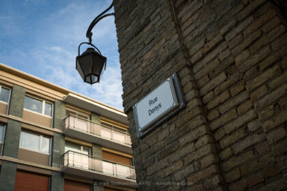 An old street sign reading Rue Denys sits fixed to a weathered brick corner in Dieppe. A traditional iron lamp hangs above the junction while a modern apartment block rises opposite. The image contrasts textures, eras, and urban change, revealing quiet layers of the town’s architecture and everyday geography today.