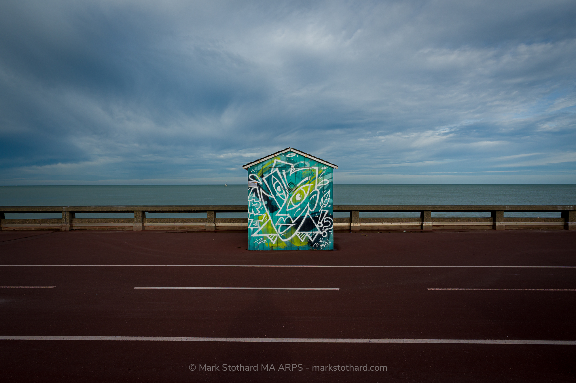 On the Dieppe esplanade a single beach hut stands between sea and road. Its painted graffiti adds bright colour against grey sky and calm water. The empty promenade and distant sailboat suggest quiet movement and waiting, a small structure holding attention in a wide coastal landscape today under soft light.