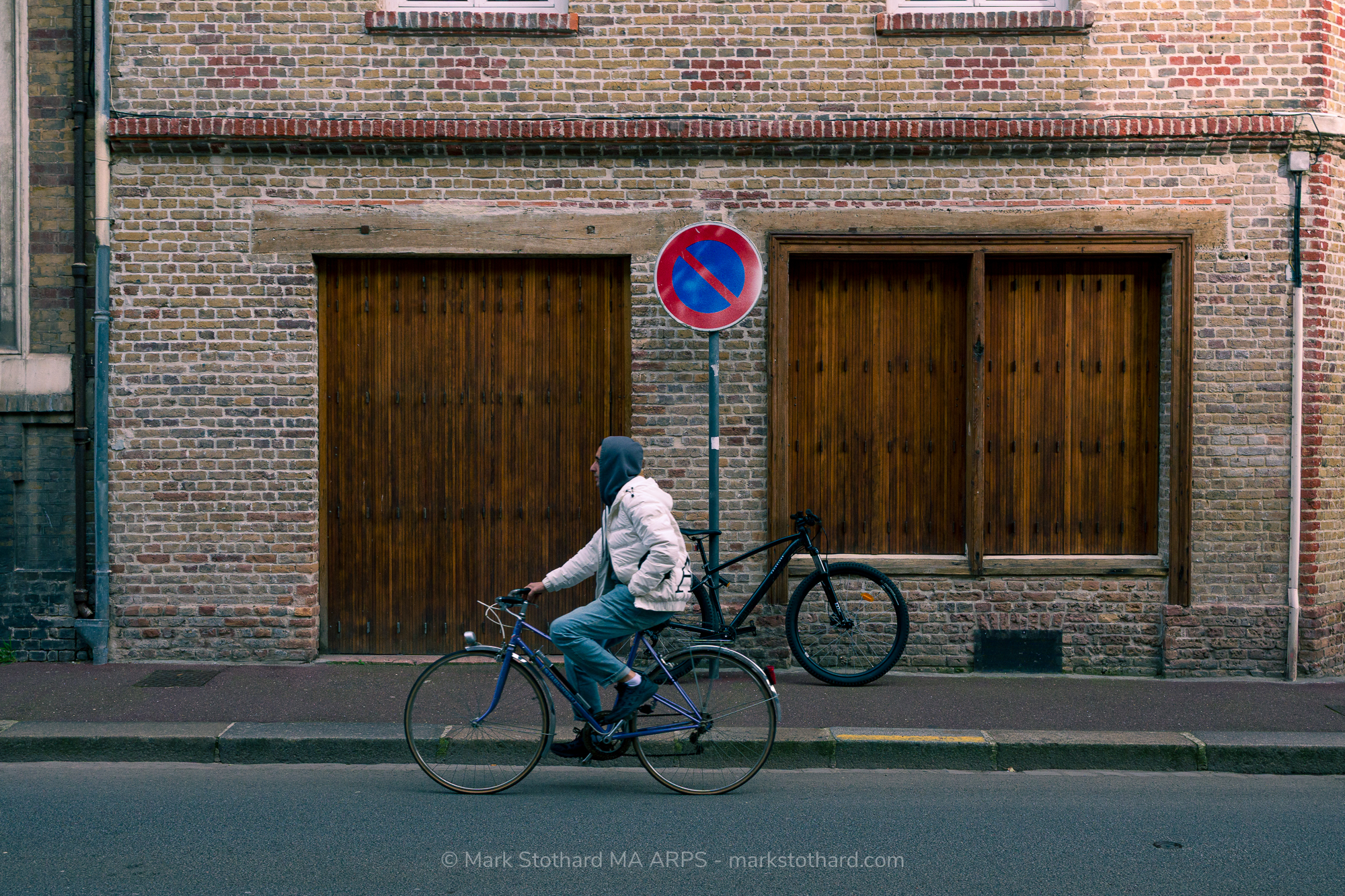 On a quiet street in Dieppe a cyclist rides past shuttered wooden doors set in a weathered brick fa&ccedil;ade. A parked bicycle waits against the wall beneath a no parking sign. The scene captures ordinary movement within still architecture, a moment of everyday rhythm in the coastal town&rsquo;s calm streets.