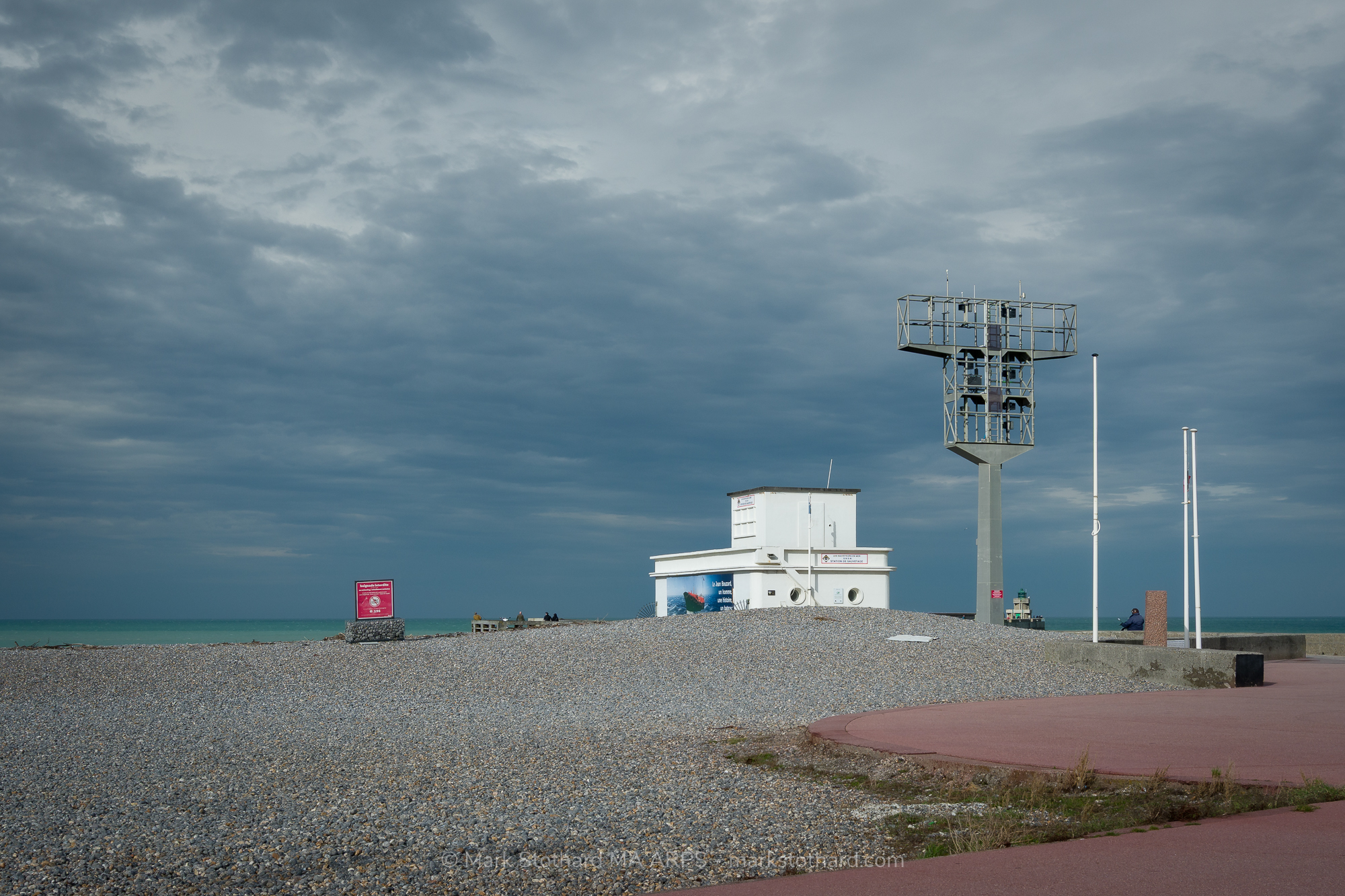Storm clouds gather above the rescue station on the Dieppe jetty. The white building and metal signal tower stand alone against the wide Channel horizon. Pebbles cover the beach in the foreground. A few distant figures watch the sea. The scene holds quiet tension between safety, weather, and open water.