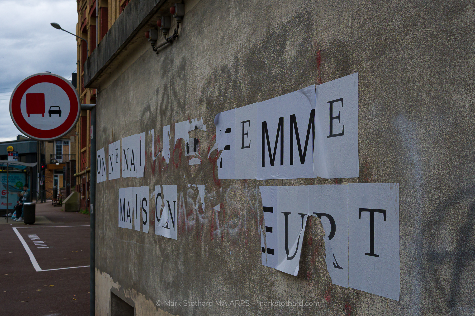 On a grey wall in Dieppe loose sheets of paper spell a message in stark black letters. The temporary posters ripple and peel against stained concrete. A traffic sign and empty street frame the scene. The image captures quiet protest fragility and the restless voice of the street today here.