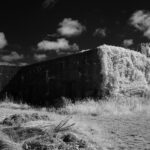This infrared photograph reveals the Kernwerk Infantry Regiment HQ in Jersey, a German WW2 command bunker, through a surreal lens. The structure’s imposing concrete form, once a symbol of occupation and defense, is now enveloped by a luminous cascade of white vegetation, as if nature itself seeks to heal the wounds of the past. The sky above is a turbulent canvas of dark clouds and fleeting light, adding a sense of drama and tension to the scene.The bunker, a Regelbau 609SK, was designed to withstand invasion, its reinforced walls and strategic placement a testament to German engineering.Today, it stands as a silent witness to history, its utilitarian purpose softened by time and the encroaching wilderness. The infrared effect transforms the landscape into an otherworldly tableau, where the stark lines of the bunker contrast with the ethereal glow of the foliage. This juxtaposition evokes a poignant reflection on the passage of time, the resilience of nature, and the enduring echoes of war. For photographers and historians, Kernwerk offers a rare opportunity to capture the intersection of conflict, architecture, and the quiet power of renewal.