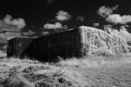 This infrared photograph reveals the Kernwerk Infantry Regiment HQ in Jersey, a German WW2 command bunker, through a surreal lens. The structure’s imposing concrete form, once a symbol of occupation and defense, is now enveloped by a luminous cascade of white vegetation, as if nature itself seeks to heal the wounds of the past. The sky above is a turbulent canvas of dark clouds and fleeting light, adding a sense of drama and tension to the scene.The bunker, a Regelbau 609SK, was designed to withstand invasion, its reinforced walls and strategic placement a testament to German engineering.Today, it stands as a silent witness to history, its utilitarian purpose softened by time and the encroaching wilderness. The infrared effect transforms the landscape into an otherworldly tableau, where the stark lines of the bunker contrast with the ethereal glow of the foliage. This juxtaposition evokes a poignant reflection on the passage of time, the resilience of nature, and the enduring echoes of war. For photographers and historians, Kernwerk offers a rare opportunity to capture the intersection of conflict, architecture, and the quiet power of renewal.